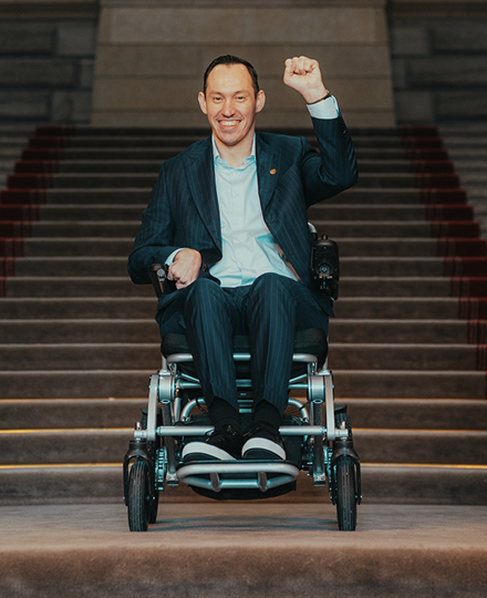 Islam Alijaj im Rollstuhl auf einer Treppe vor dem Bundeshaus, die Faust erhoben.